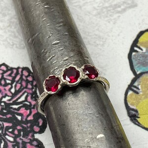 May include: A silver ring featuring three round, deep red gemstones. The gemstones are set in a floral design. The ring is displayed on a metal rod, highlighting its delicate details and craftsmanship.