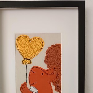 May include: A framed artwork featuring a red stitched animal holding a yellow heart-shaped balloon. The animal is stitched with a white outline and has a white stitched collar. The background is a light beige color.