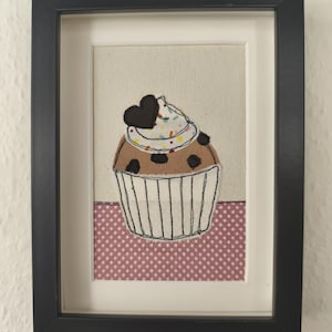 May include: Framed artwork featuring a cupcake design. The cupcake has a brown base with chocolate chips, white frosting, and a black heart on top. The artwork is framed in a black frame and set against a neutral background.