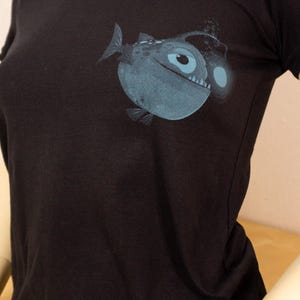 Peut inclure: T-shirt noir avec un motif d'un poisson pêcheur bleu. L'illustration comprend le corps du poisson, son œil et un leurre lumineux. Le t-shirt est présenté sur un mannequin.