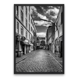 May include: A black and white photograph of a narrow cobblestone street in a European city. The street is lined with buildings on both sides, and there are a few people walking in the distance. The sky is cloudy, and the street is empty except for a bicycle parked on the right side.