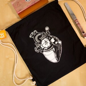 Könnte beinhalten: Schwarze Kordeltasche mit einem weißen Aufdruck eines Steampunk-Herzens. Die Tasche ist aus Canvas und hat einen weißen Kordelzug.