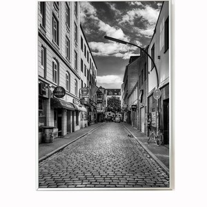 May include: A black and white photo of a cobblestone street in a city. The street is lined with buildings on both sides, and there is a bicycle parked on the right side of the street.