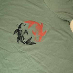 K&ouml;nnte beinhalten: Salbeigr&uuml;nes T-Shirt mit einer Grafik von zwei Koi-Karpfen, einer schwarzen und einer roten, in einem Yin-Yang-Design. Die Fische sind stilisiert mit detaillierten Flossen und Schuppen. Ein l&auml;ssiges Oberteil f&uuml;r den Alltag.