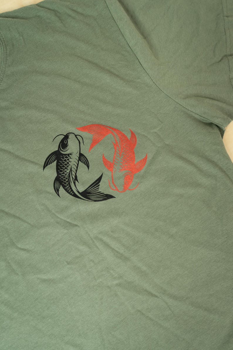 K&ouml;nnte beinhalten: Salbeigr&uuml;nes T-Shirt mit einer Grafik von zwei Koi-Karpfen, einer schwarzen und einer roten, in einem Yin-Yang-Design. Die Fische sind stilisiert mit detaillierten Flossen und Schuppen. Ein l&auml;ssiges Oberteil f&uuml;r den Alltag.