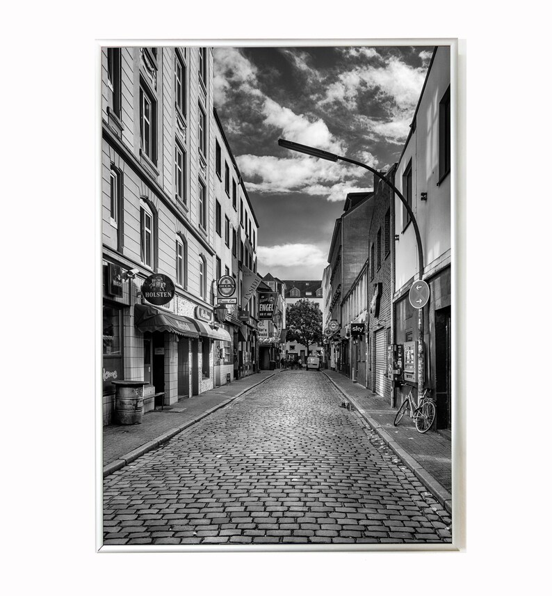 May include: A black and white photo of a cobblestone street in a city. The street is lined with buildings on both sides, and there is a bicycle parked on the right side of the street.