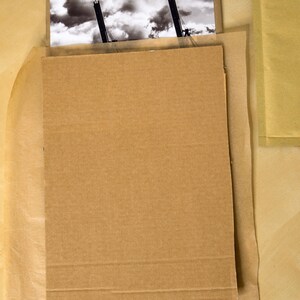 May include: A black and white photograph of two cranes against a cloudy sky is protected by a cardboard backing and brown paper wrapping.