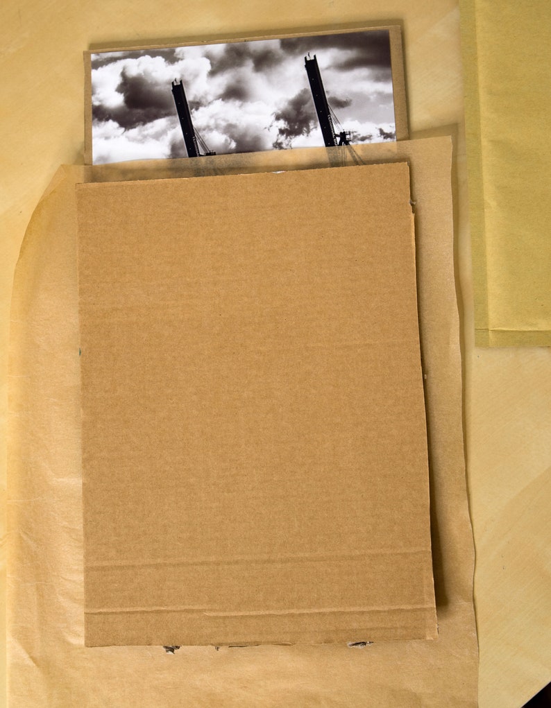 May include: A black and white photograph of two cranes against a cloudy sky is protected by a cardboard backing and brown paper wrapping.