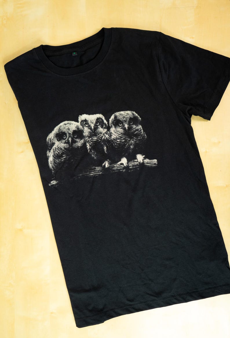 May include: A black short-sleeved t-shirt featuring a white graphic print of three owls perched together on a branch. The owls are depicted in a detailed, sketch-like style, centered on the chest area of the garment.
