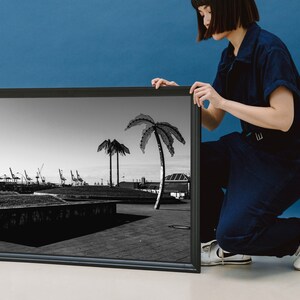 May include: A black and white framed photograph of a cityscape with palm trees in the foreground. The photograph is being held by a person wearing a blue jumpsuit.