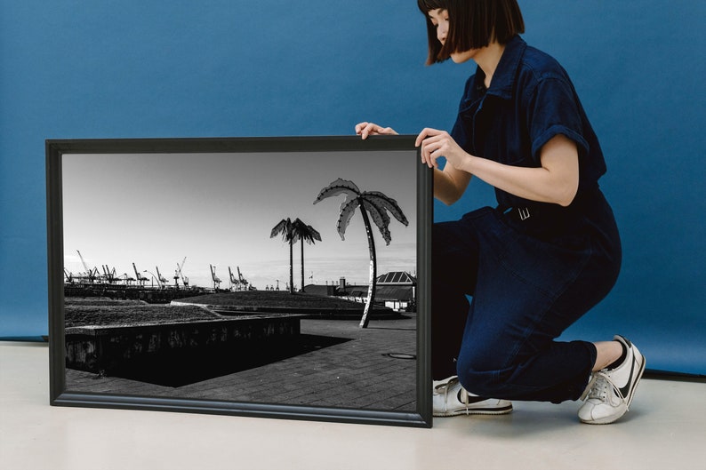 May include: A black and white framed photograph of a cityscape with palm trees in the foreground. The photograph is being held by a person wearing a blue jumpsuit.