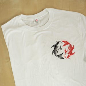 K&ouml;nnte beinhalten: Wei&szlig;es T-Shirt mit einem schwarzen und roten Koi-Fisch-Design. Die Grafik zeigt zwei Fische in einem kreisf&ouml;rmigen Muster, einen schwarzen und einen roten, die Ausgewogenheit symbolisieren. Das Shirt besteht aus einem weichen, bequemen Material.