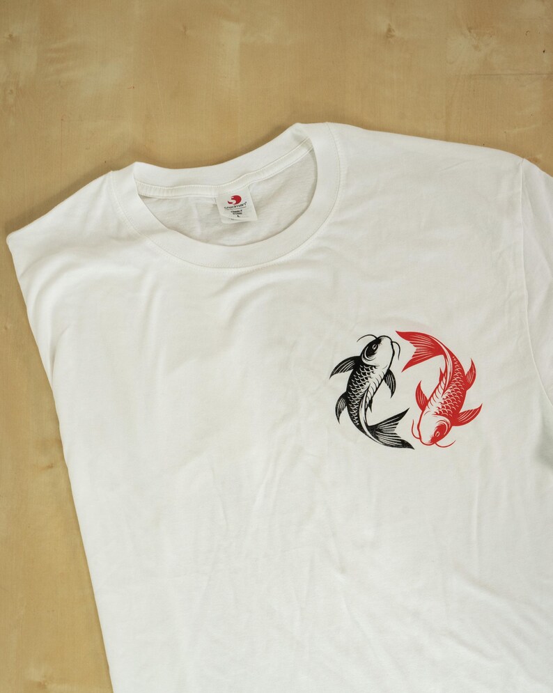 K&ouml;nnte beinhalten: Wei&szlig;es T-Shirt mit einem schwarzen und roten Koi-Fisch-Design. Die Grafik zeigt zwei Fische in einem kreisf&ouml;rmigen Muster, einen schwarzen und einen roten, die Ausgewogenheit symbolisieren. Das Shirt besteht aus einem weichen, bequemen Material.