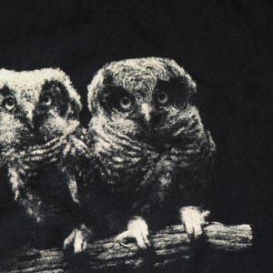 May include: A black fabric garment featuring a high-contrast, white graphic print of two owls perched side-by-side on a branch. The owls are depicted with detailed feathers and large, forward-facing eyes, set against a solid black background.