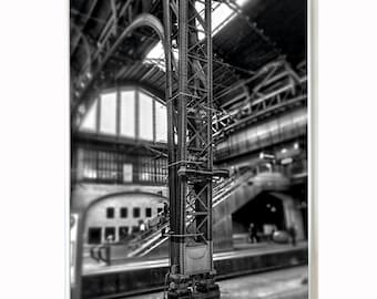 May include: A black and white photo of a large metal structure inside a building. The structure is made of beams and supports, and it appears to be a part of a crane or other industrial equipment. The photo is taken from a low angle, looking up at the structure.