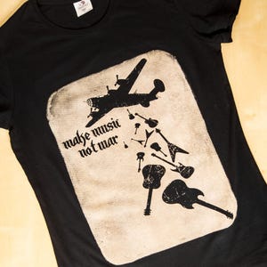 Könnte beinhalten: Schwarzes T-Shirt mit einem Vintage-Grafikdruck eines Flugzeugs, das Gitarren mit dem Text "make music not war" abwirft.