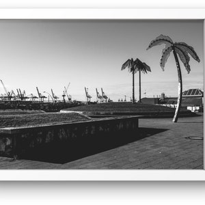 May include: A black and white photograph of a concrete plaza with two palm trees in the foreground. The palm trees are made of metal and have a modern, abstract design. In the background, there are several industrial cranes and buildings.