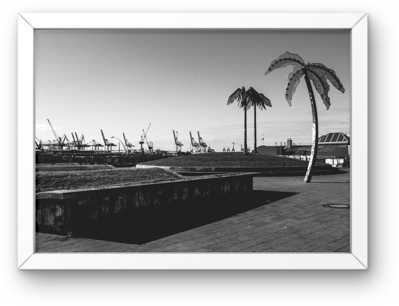 May include: A black and white photograph of a concrete plaza with two palm trees in the foreground. The palm trees are made of metal and have a modern, abstract design. In the background, there are several industrial cranes and buildings.