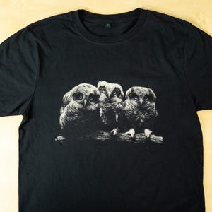 Black t-shirt laid flat on a light wooden surface featuring a monochrome print of three owls perched on a branch in a dark vintage wildlife style.