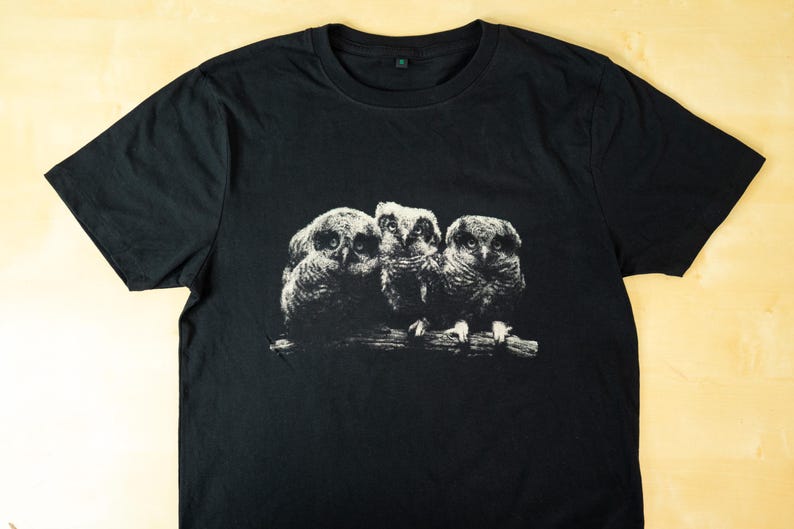 Black t-shirt laid flat on a light wooden surface featuring a monochrome print of three owls perched on a branch in a dark vintage wildlife style.