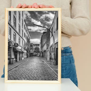 May include: Black and white photograph of a cobblestone street in a European city. The street is lined with buildings on both sides, and there are trees and streetlights in the background.