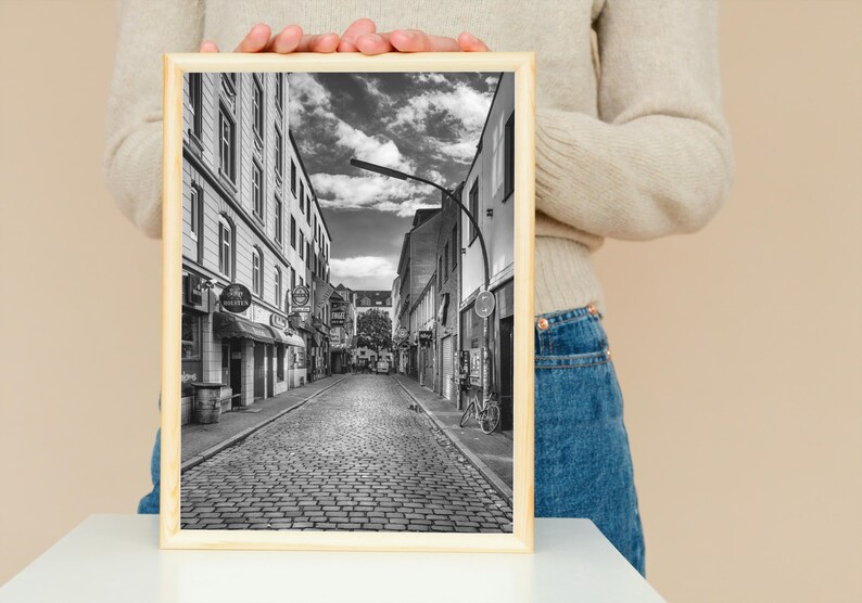 May include: Black and white photograph of a cobblestone street in a European city. The street is lined with buildings on both sides, and there are trees and streetlights in the background.