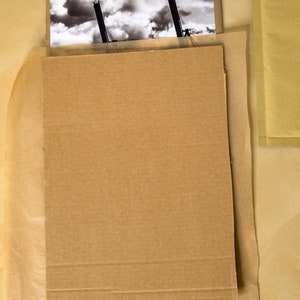 Op de afbeelding: Een zwart-witfoto van twee kranen tegen een bewolkte lucht is beschermd door een kartonnen achterkant en bruine verpakking.