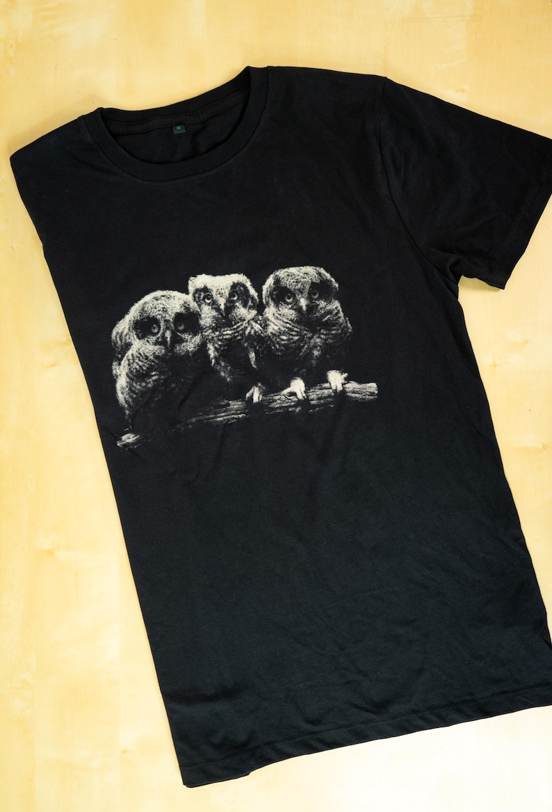 Black t-shirt laid flat on a light wooden surface featuring a monochrome print of three owls perched on a branch in a dark vintage wildlife style.