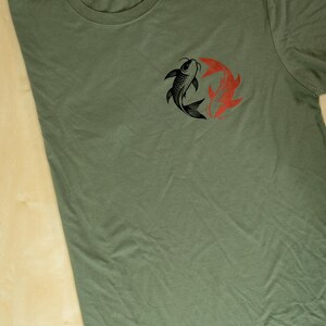 K&ouml;nnte beinhalten: Olivgr&uuml;nes T-Shirt mit einem kreisf&ouml;rmigen Design aus zwei Koi-Karpfen, einem schwarzen und einem roten. Die Fische sind im Yin-Yang-Stil angeordnet. Das T-Shirt besteht aus einem weichen Material und hat einen Rundhalsausschnitt.
