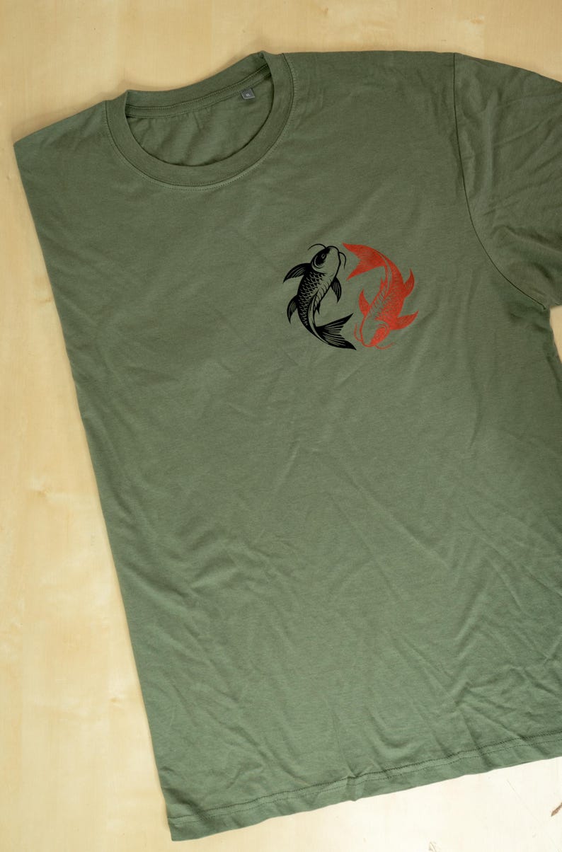 K&ouml;nnte beinhalten: Olivgr&uuml;nes T-Shirt mit einem kreisf&ouml;rmigen Design aus zwei Koi-Karpfen, einem schwarzen und einem roten. Die Fische sind im Yin-Yang-Stil angeordnet. Das T-Shirt besteht aus einem weichen Material und hat einen Rundhalsausschnitt.