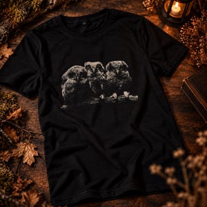 May include: A black short-sleeved t-shirt featuring a detailed white graphic print of three owls perched together on a branch. The design is centered on the chest of the shirt, showcasing the textured feathers and distinct facial features of the birds.