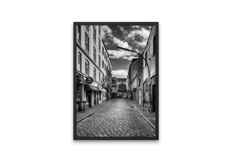May include: A black and white photograph of a narrow cobblestone street in a European city. The street is lined with buildings on both sides, and there are a few people walking in the distance. The sky is cloudy, and the street is empty except for a bicycle parked on the right side.