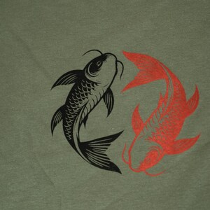 K&ouml;nnte beinhalten: Olivgr&uuml;nes T-Shirt mit einem Design aus schwarzen und roten Koi-Fischen. Die Fische sind kreisf&ouml;rmig angeordnet, einer schwarz und einer rot. Das Design ist mittig auf dem T-Shirt platziert.