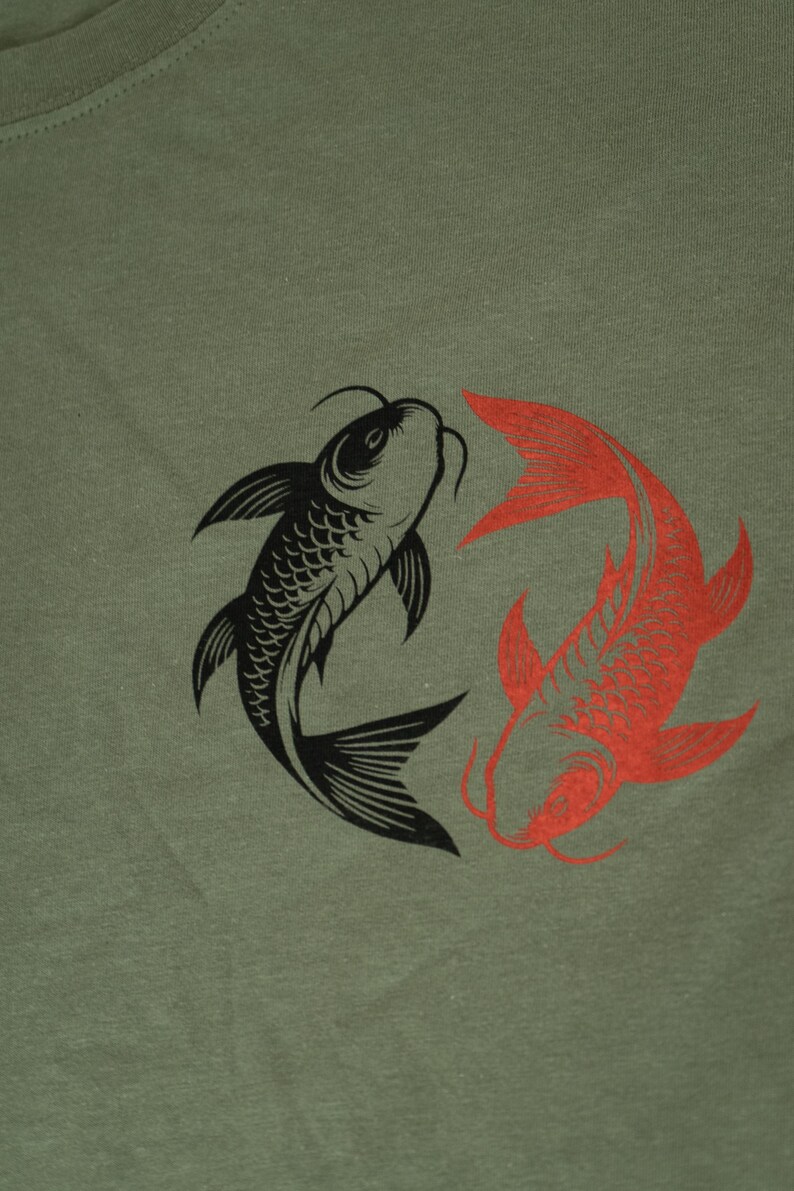K&ouml;nnte beinhalten: Olivgr&uuml;nes T-Shirt mit einem Design aus schwarzen und roten Koi-Fischen. Die Fische sind kreisf&ouml;rmig angeordnet, einer schwarz und einer rot. Das Design ist mittig auf dem T-Shirt platziert.