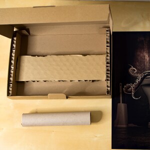 Puede incluir: Una caja de cart&oacute;n marr&oacute;n con un inserto de cart&oacute;n corrugado. La caja est&aacute; abierta y el inserto es visible. Un tubo de cart&oacute;n marr&oacute;n est&aacute; al lado de la caja. Una fotograf&iacute;a en blanco y negro de un pulpo envuelto alrededor de un inodoro es visible en el fondo.