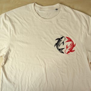 K&ouml;nnte beinhalten: Cremefarbenes T-Shirt mit einem Design aus schwarzen und roten Koi-Fischen. Die Fische sind in einem kreisf&ouml;rmigen Muster angeordnet, das Ausgeglichenheit symbolisiert. Das Shirt hat einen Rundhalsausschnitt und kurze &Auml;rmel.