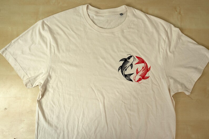 K&ouml;nnte beinhalten: Cremefarbenes T-Shirt mit einem Design aus schwarzen und roten Koi-Fischen. Die Fische sind in einem kreisf&ouml;rmigen Muster angeordnet, das Ausgeglichenheit symbolisiert. Das Shirt hat einen Rundhalsausschnitt und kurze &Auml;rmel.