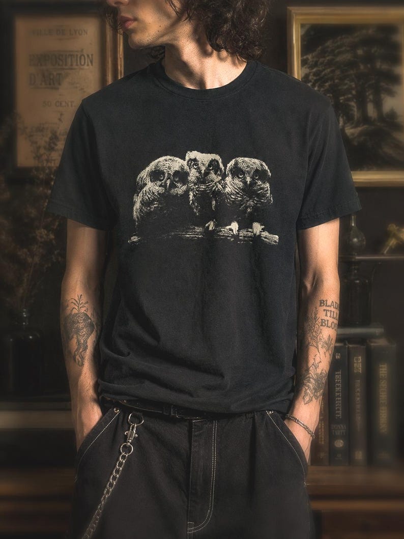 Black gothic owl t-shirt worn by a person in a dark academia styled room with vintage books, moody lighting, and monochrome owl graphic print.