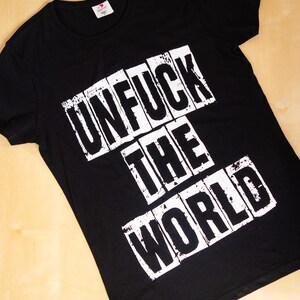 Könnte beinhalten: Schwarzes T-Shirt mit dem weißen Aufdruck "UNFUCK THE WORLD" in Blockbuchstaben. Der Text ist in drei Zeilen angeordnet. Das T-Shirt hat einen Rundhalsausschnitt und ist aus weichem Material.