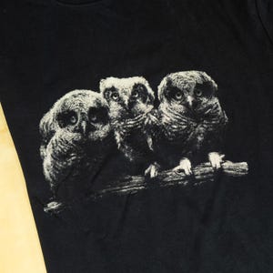 May include: A black t-shirt featuring a graphic print of three owls perched together on a branch. The owls are rendered in a textured, off-white and gray monochromatic style, showing detailed feathers and large, forward-facing eyes.