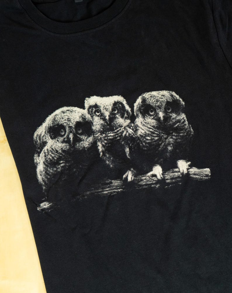 May include: A black t-shirt featuring a graphic print of three owls perched together on a branch. The owls are rendered in a textured, off-white and gray monochromatic style, showing detailed feathers and large, forward-facing eyes.