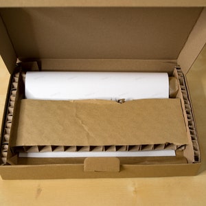 May include: A brown cardboard box with its lid open, revealing a white rolled-up item inside. The box is filled with brown packing peanuts for protection.