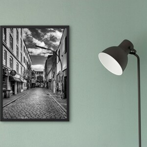 May include: Black and white photograph of a cobblestone street in a city. The street is lined with buildings on both sides. The buildings are tall and have many windows. There is a street lamp in the middle of the street.