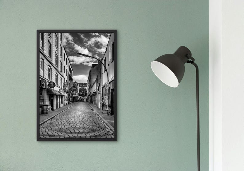 May include: Black and white photograph of a cobblestone street in a city. The street is lined with buildings on both sides. The buildings are tall and have many windows. There is a street lamp in the middle of the street.