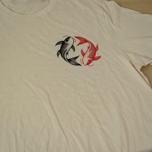 K&ouml;nnte beinhalten: Cremefarbenes T-Shirt mit einem Design aus schwarzen und roten Koi-Karpfen. Die Fische sind kreisf&ouml;rmig angeordnet, einer schwarz und einer rot. Das Shirt hat einen Rundhalsausschnitt und kurze &Auml;rmel.