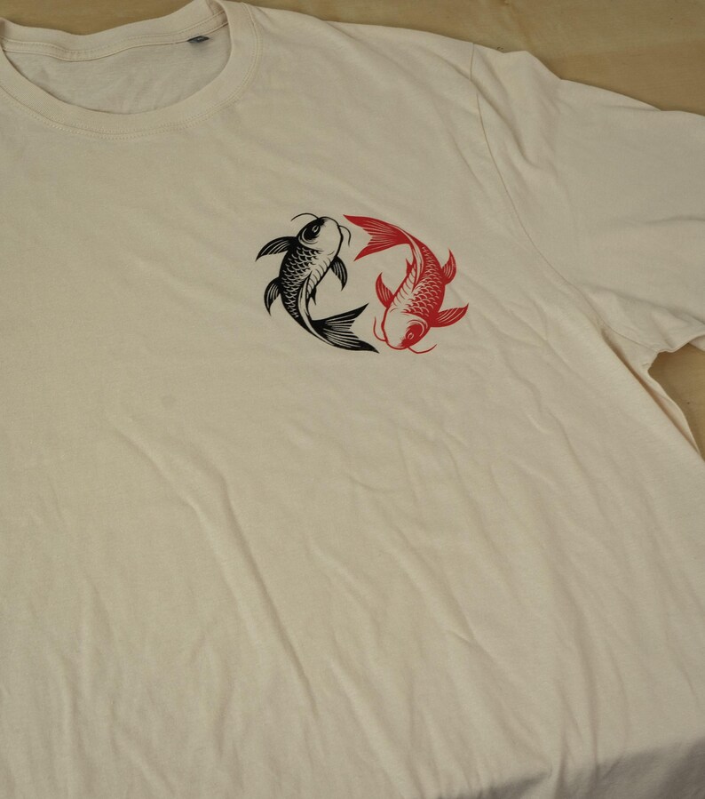K&ouml;nnte beinhalten: Cremefarbenes T-Shirt mit einem Design aus schwarzen und roten Koi-Karpfen. Die Fische sind kreisf&ouml;rmig angeordnet, einer schwarz und einer rot. Das Shirt hat einen Rundhalsausschnitt und kurze &Auml;rmel.