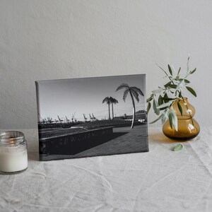 May include: Black and white photograph of a harbor scene with palm trees and cranes printed on a canvas. The canvas is sitting on a white linen tablecloth with a lit candle and a vase with green branches.
