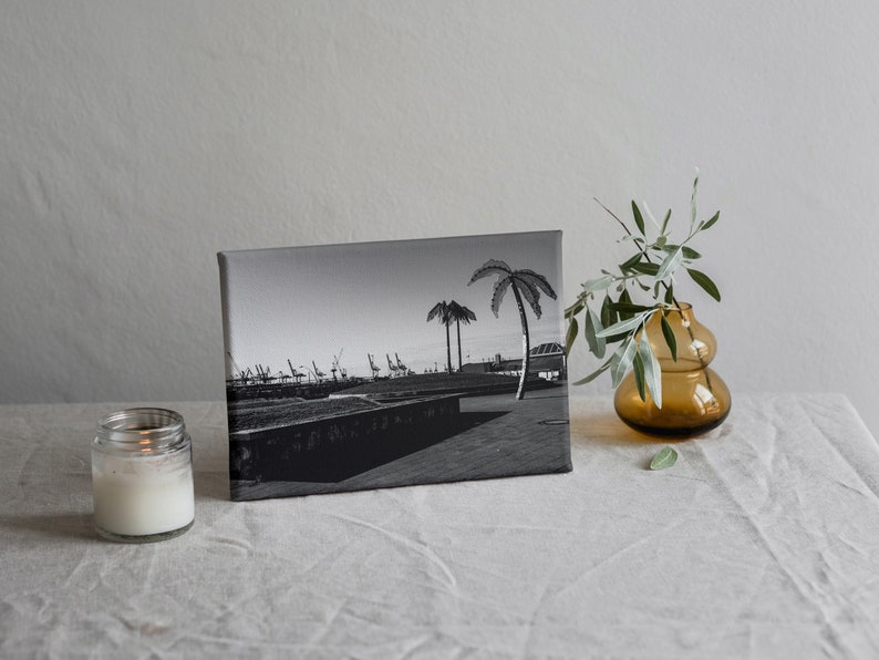 May include: Black and white photograph of a harbor scene with palm trees and cranes printed on a canvas. The canvas is sitting on a white linen tablecloth with a lit candle and a vase with green branches.
