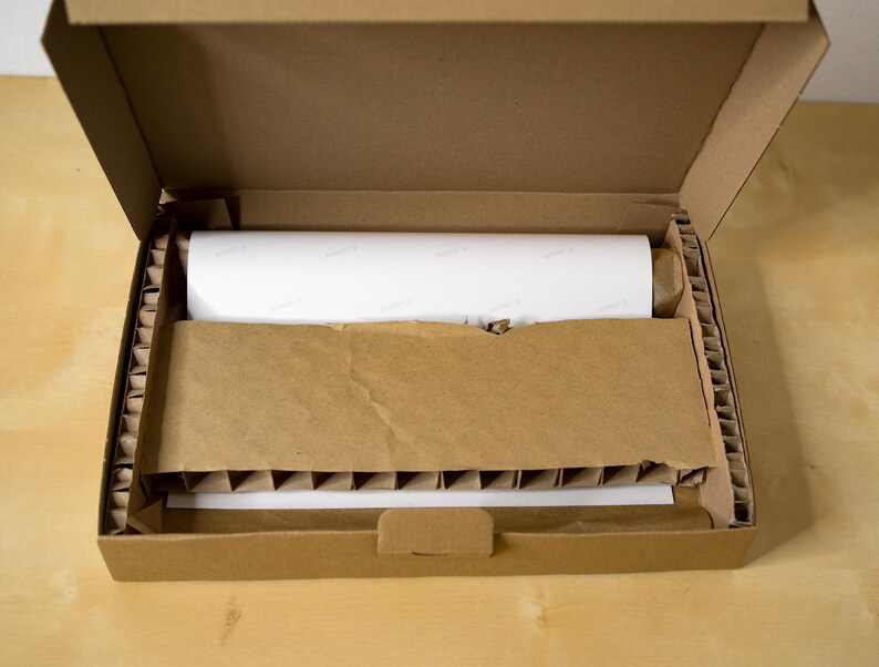 Puede incluir: Una caja de cart&oacute;n marr&oacute;n con la tapa abierta, revelando un art&iacute;culo blanco enrollado en el interior. La caja est&aacute; llena de cacahuetes de embalaje marrones para protecci&oacute;n.