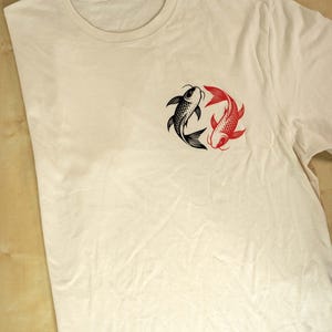 K&ouml;nnte beinhalten: Cremefarbenes T-Shirt mit einem schwarzen und roten Koi-Fisch-Design. Die Fische sind in einem kreisf&ouml;rmigen Muster angeordnet, wobei sich ein schwarzer und ein roter Fisch gegen&uuml;berstehen. Das Shirt hat einen Rundhalsausschnitt.